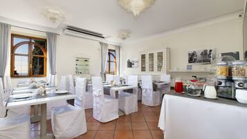 Zona de menjador amb taules preparades per a una refecció, cadires blanques i un aparador amb diferents aliments.