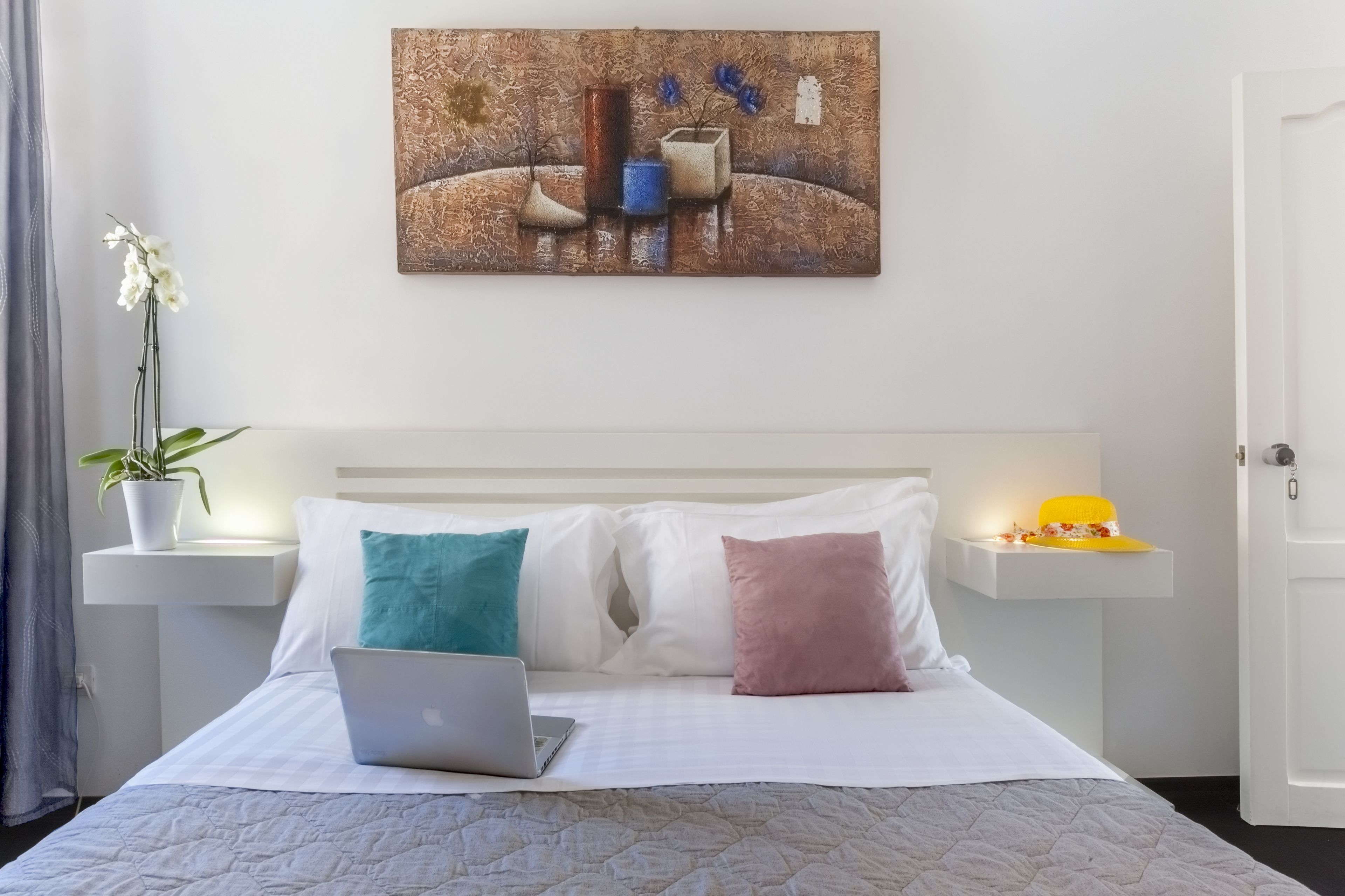Una cama bien hecha con una computadora portátil, almohadas blancas y un cuadro en la pared.