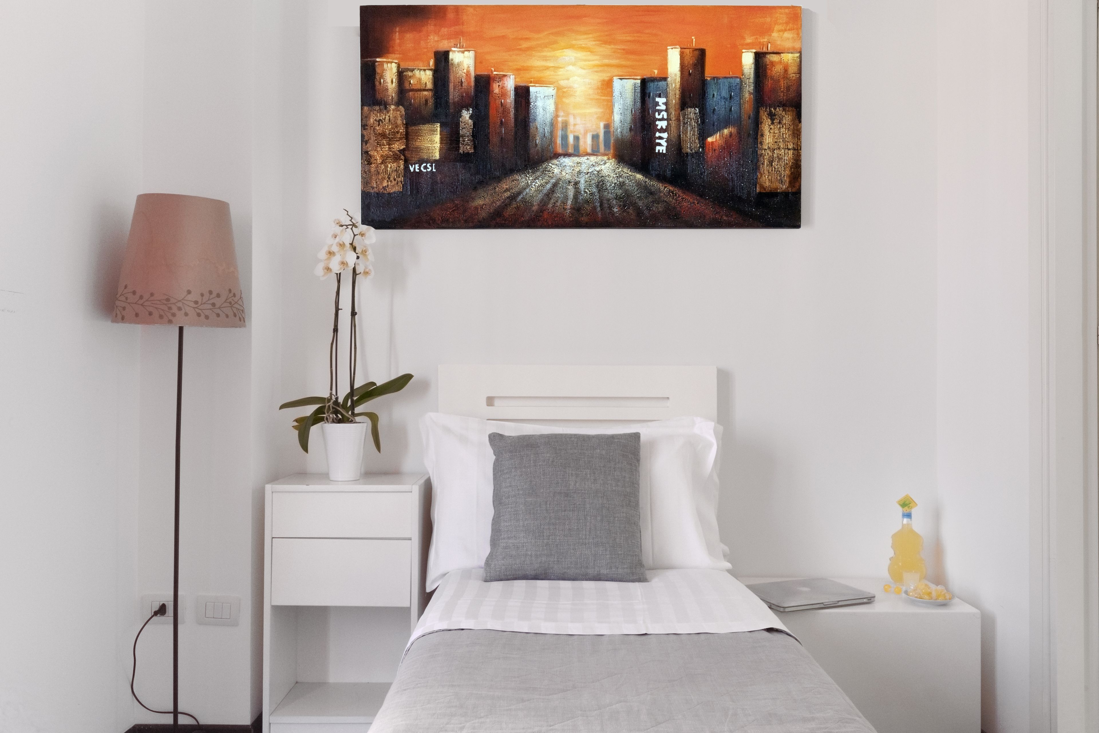 A bedroom with a bed, a nightstand, a lamp, a potted plant, and a painting of a cityscape.