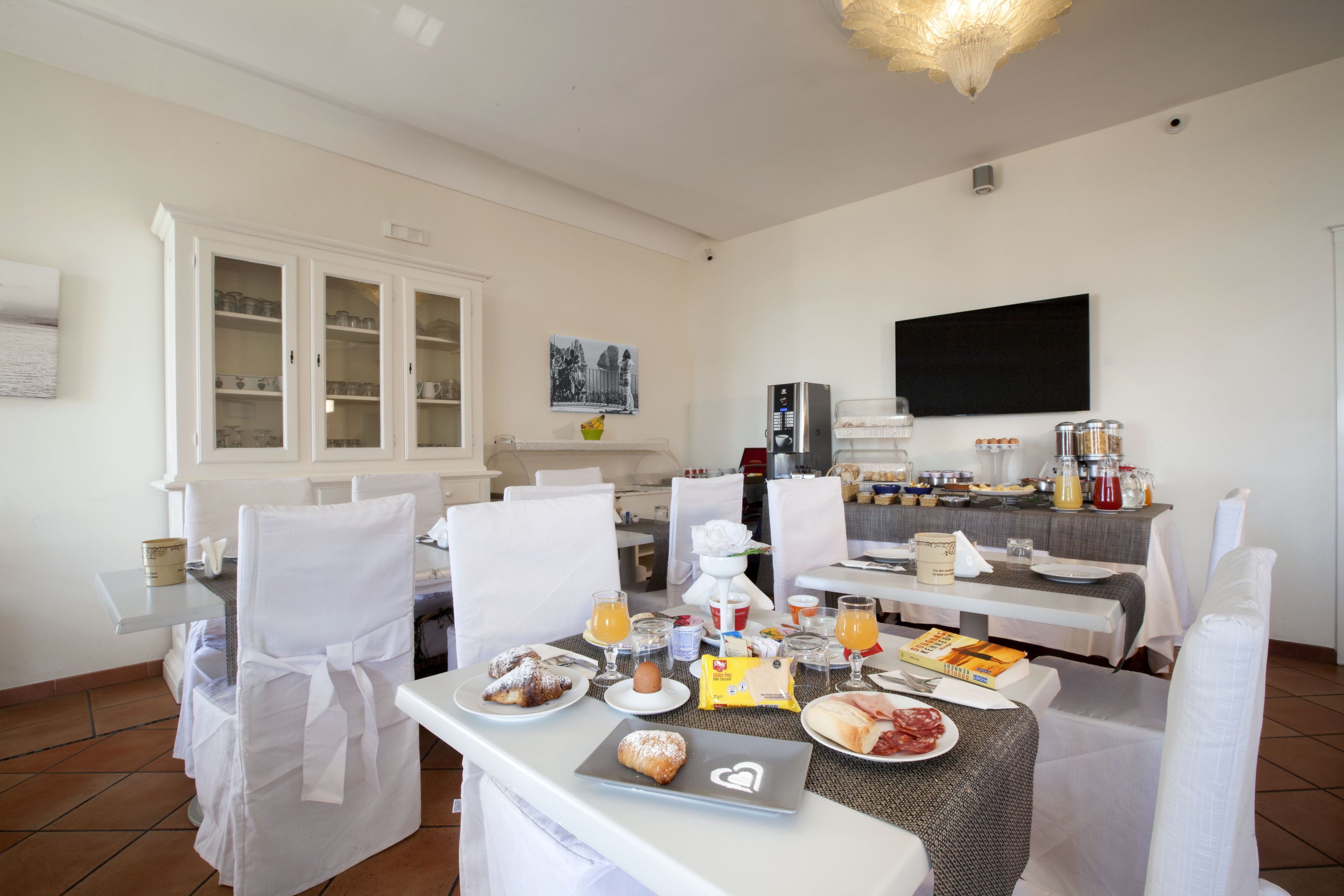 A breakfast setup with a table set for two, including plates with pastries, a muffin, and a croissant. There are two chairs with white covers, a white table, and a white cabinet in the background.