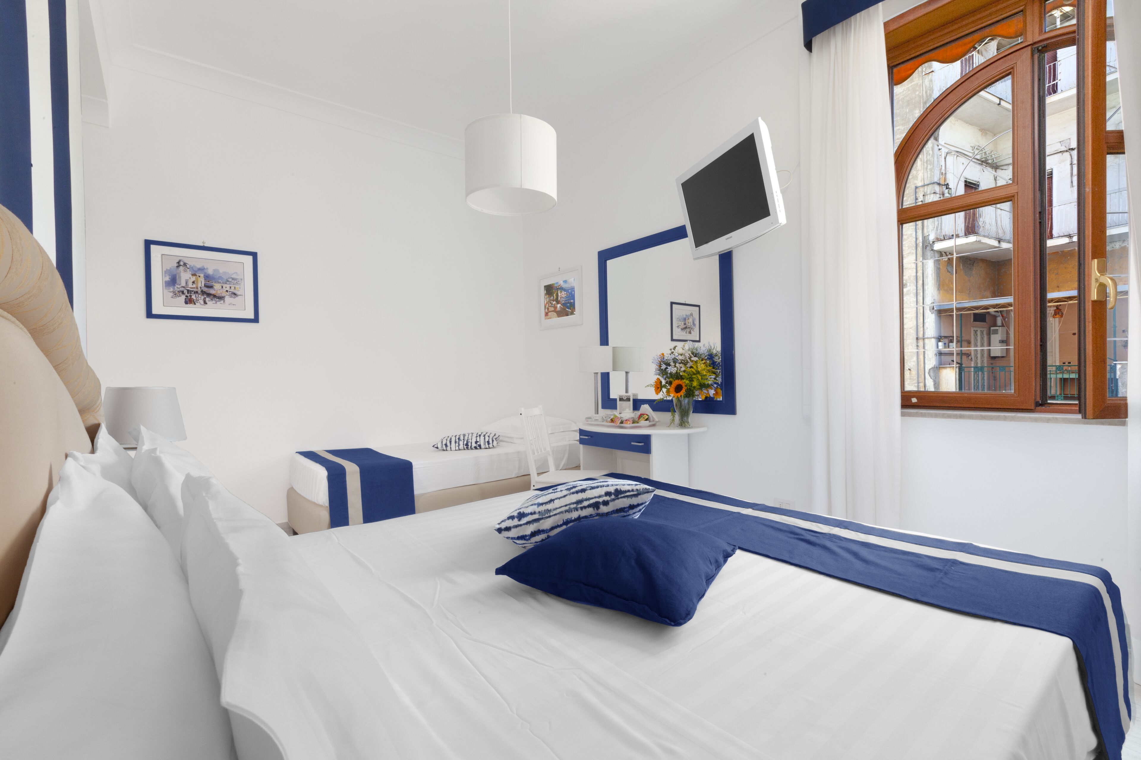 A hotel room with a bed, bedside table, lamp, TV, and a window with a view of buildings.