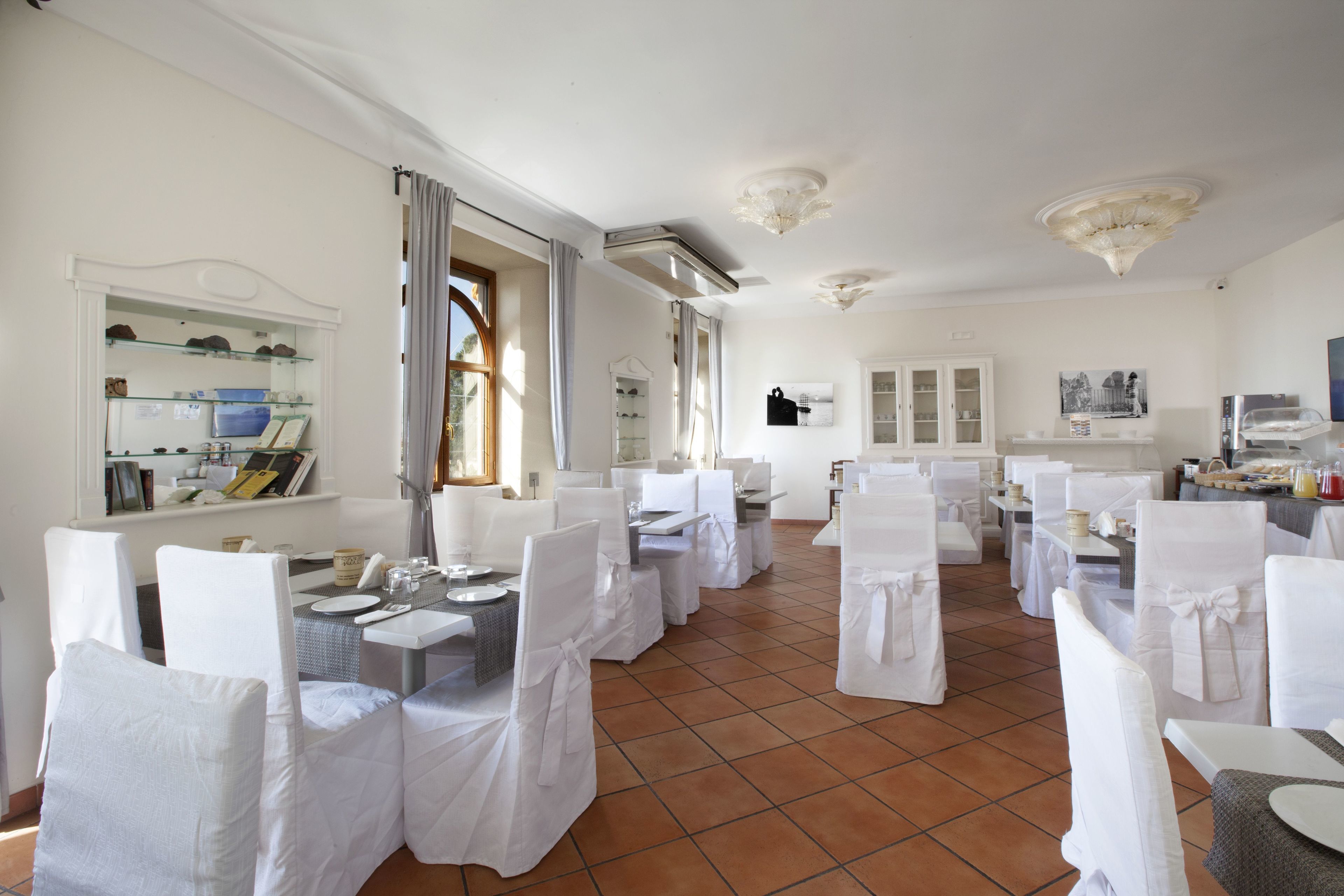 Un comedor con sillas y mesas blancas, una zona de cocina con mesas de buffet, y un amplio ventanal con cortinas.