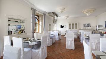 Un comedor con sillas y mesas blancas, una zona de cocina con mesas de buffet, y un amplio ventanal con cortinas.