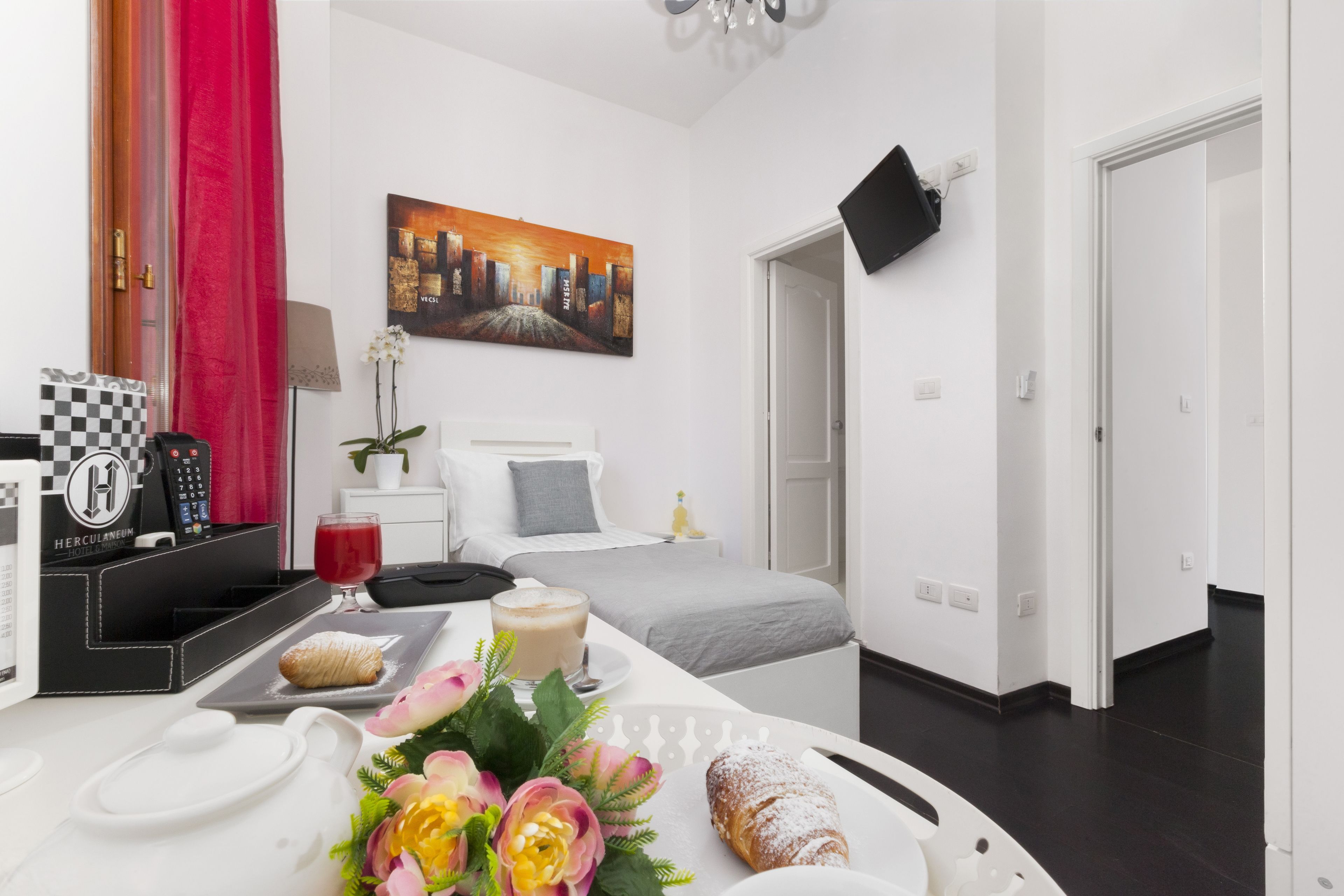 Ein modernes Hotelzimmer mit Bett, Schreibtisch mit Laptop, einer Kaffeemaschine und einer Frühstücksauslage mit Gebäck und Blumen.