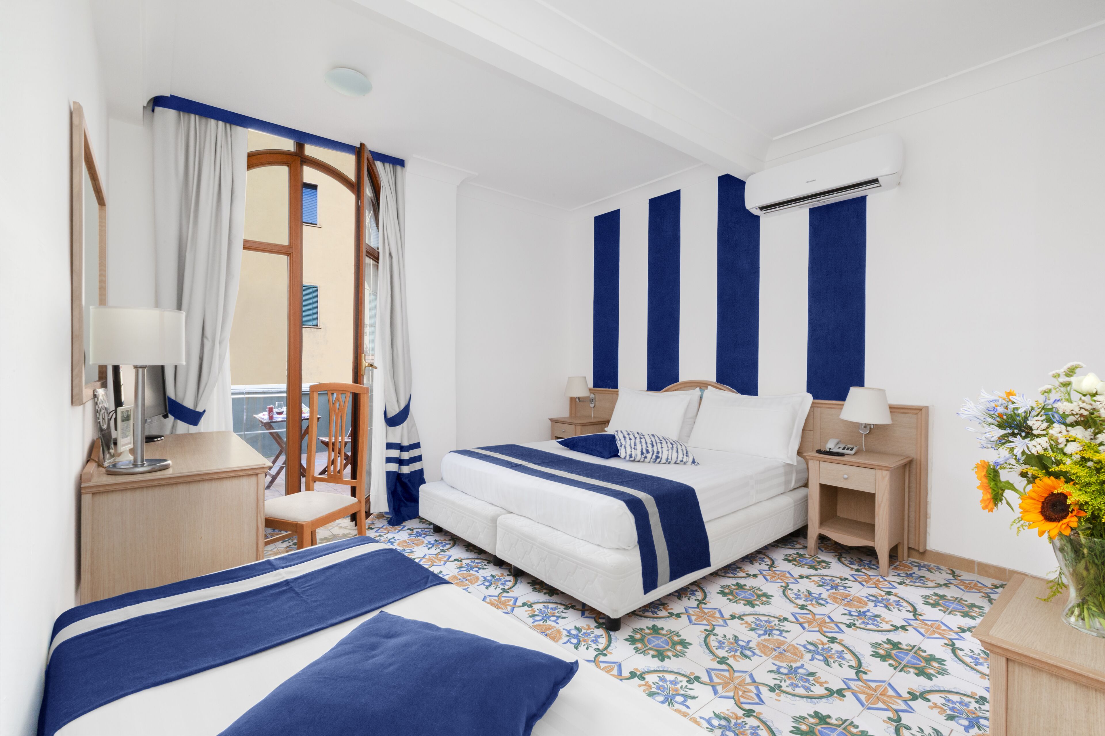 Une chambre d’hôtel avec un lit, des tables de chevet, une chaise, une fenêtre avec des rideaux et un vase avec des fleurs.