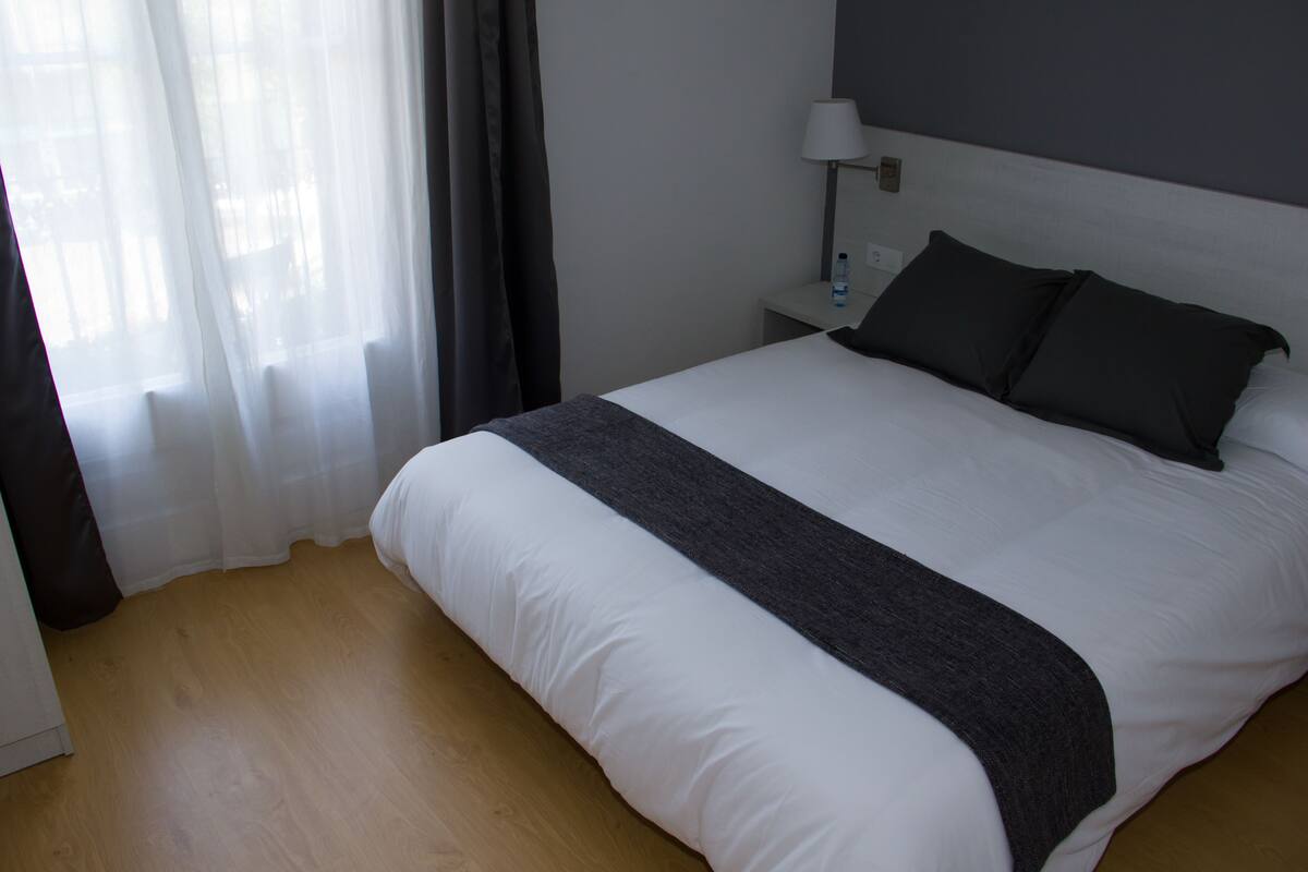 A neatly made bed with a white duvet and dark pillows, a bedside table with a lamp, and a window with sheer curtains.