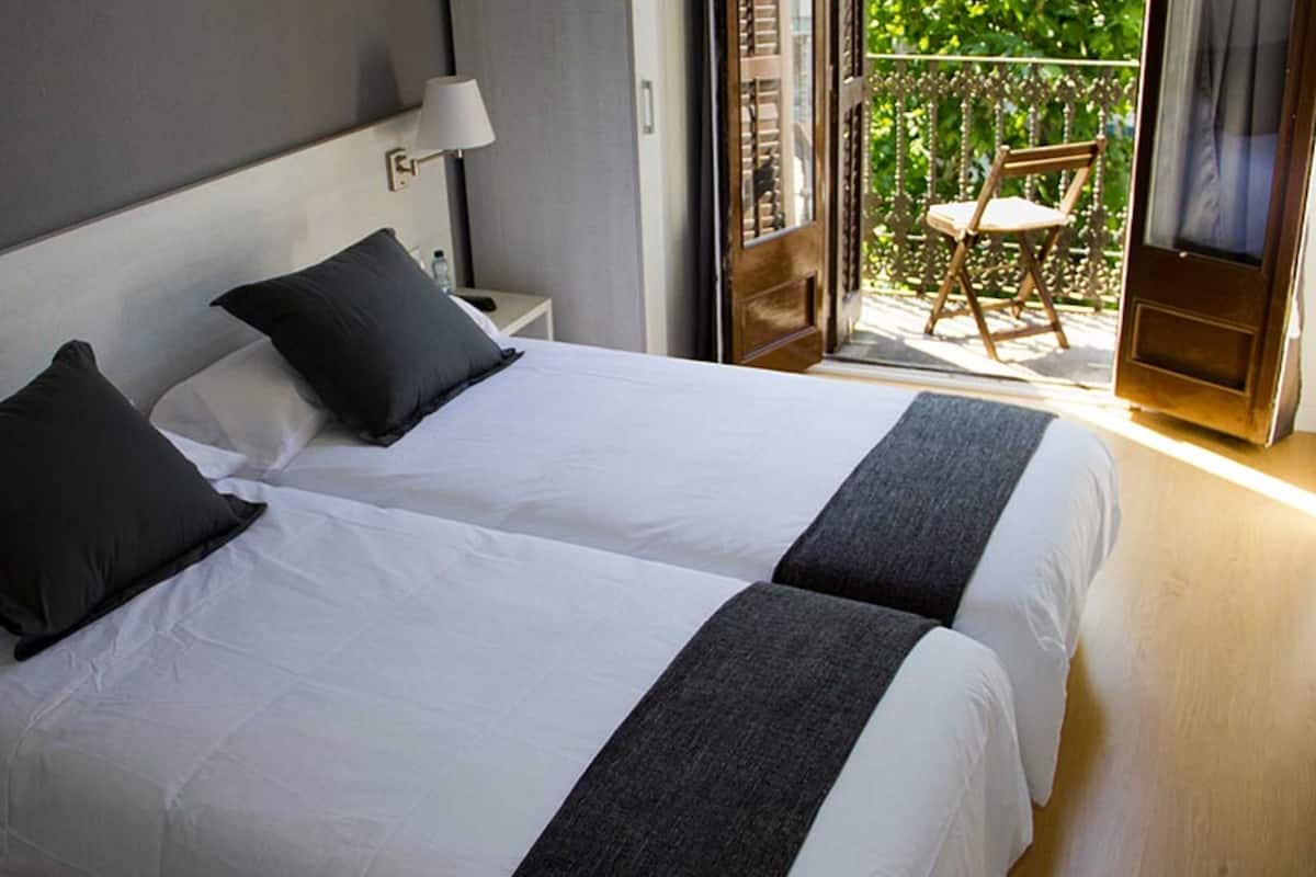 A hotel room with two beds, a nightstand with a lamp, and a balcony with a chair.