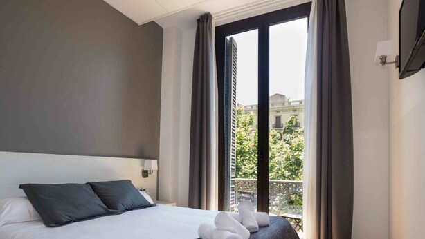 A hotel room with a large bed, grey curtains, and a view of the city through the window. - Hostalin Diputacion (Barcelona)