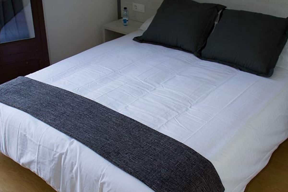 A neatly made bed with a white duvet and two dark pillows, a bedside table with a lamp, and a water bottle.
