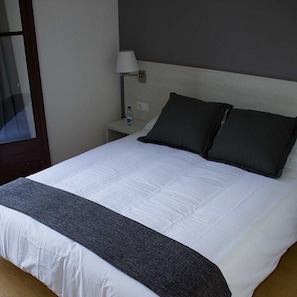 A neatly made bed with a white duvet and two dark pillows, a bedside table with a lamp, and a water bottle. - Hostalin Diputacion (Barcelona)