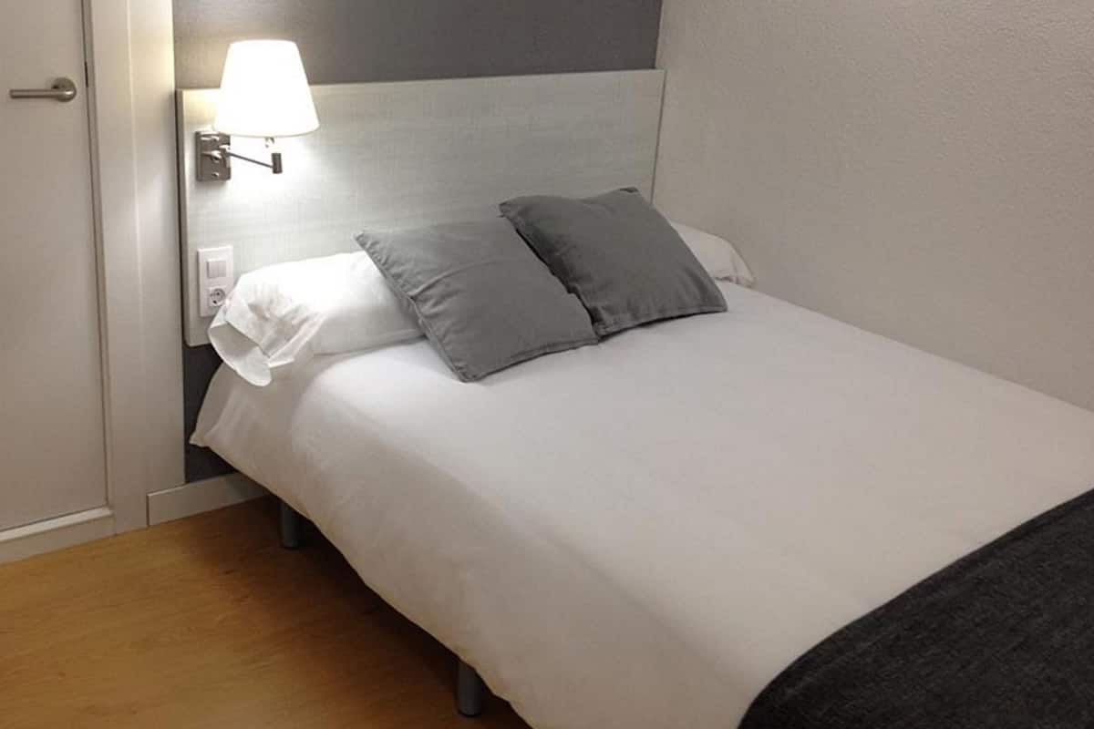 A neatly made bed with white and gray linens, a bedside lamp, and a wooden floor.