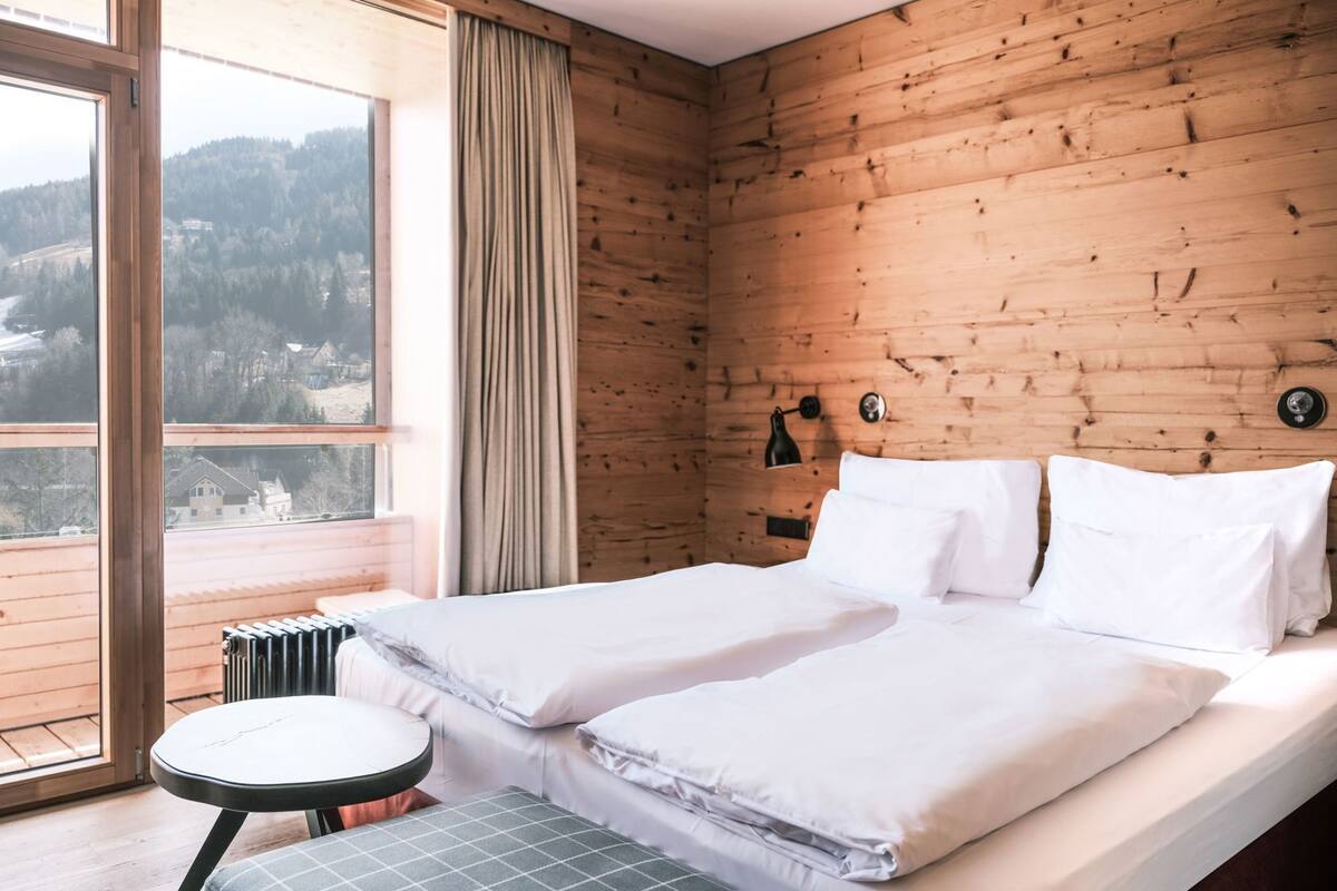 Een kamer met een houten wand, een bed met witte beddengoed, een kleine tafel en uitzicht op een bergachtig landschap.