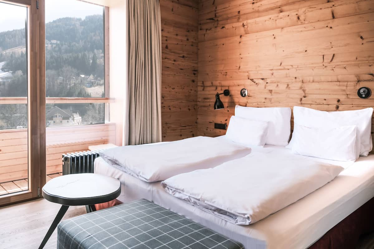 Hotelkamer met een houten wand, een bed met witte lakens, een klein rond tafeltje en uitzicht op een bebost gebied door een raam.