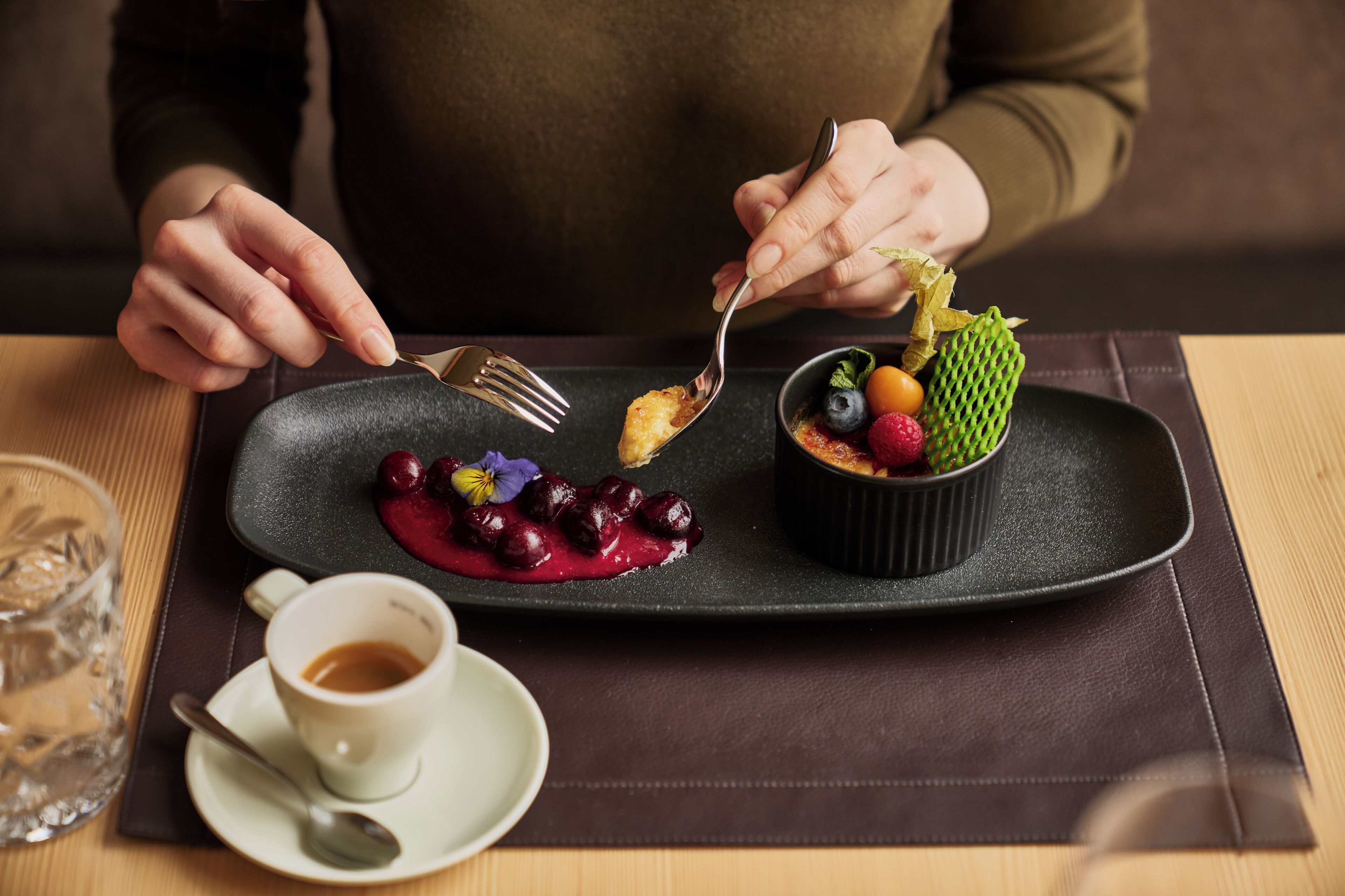 A dessert plate with a variety of fruits and a small bowl on a dark plate, a cup of coffee, and a glass of water on a table.