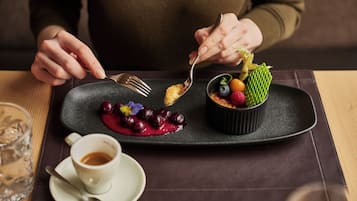 A dessert plate with a variety of fruits and a small bowl on a dark plate, a cup of coffee, and a glass of water on a table.