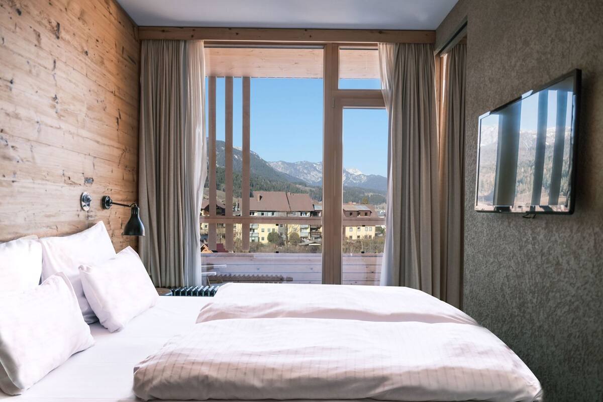 Een hotelkamer met een groot bed, een aan de muur bevestigde televisie en uitzicht op bergen door het raam.