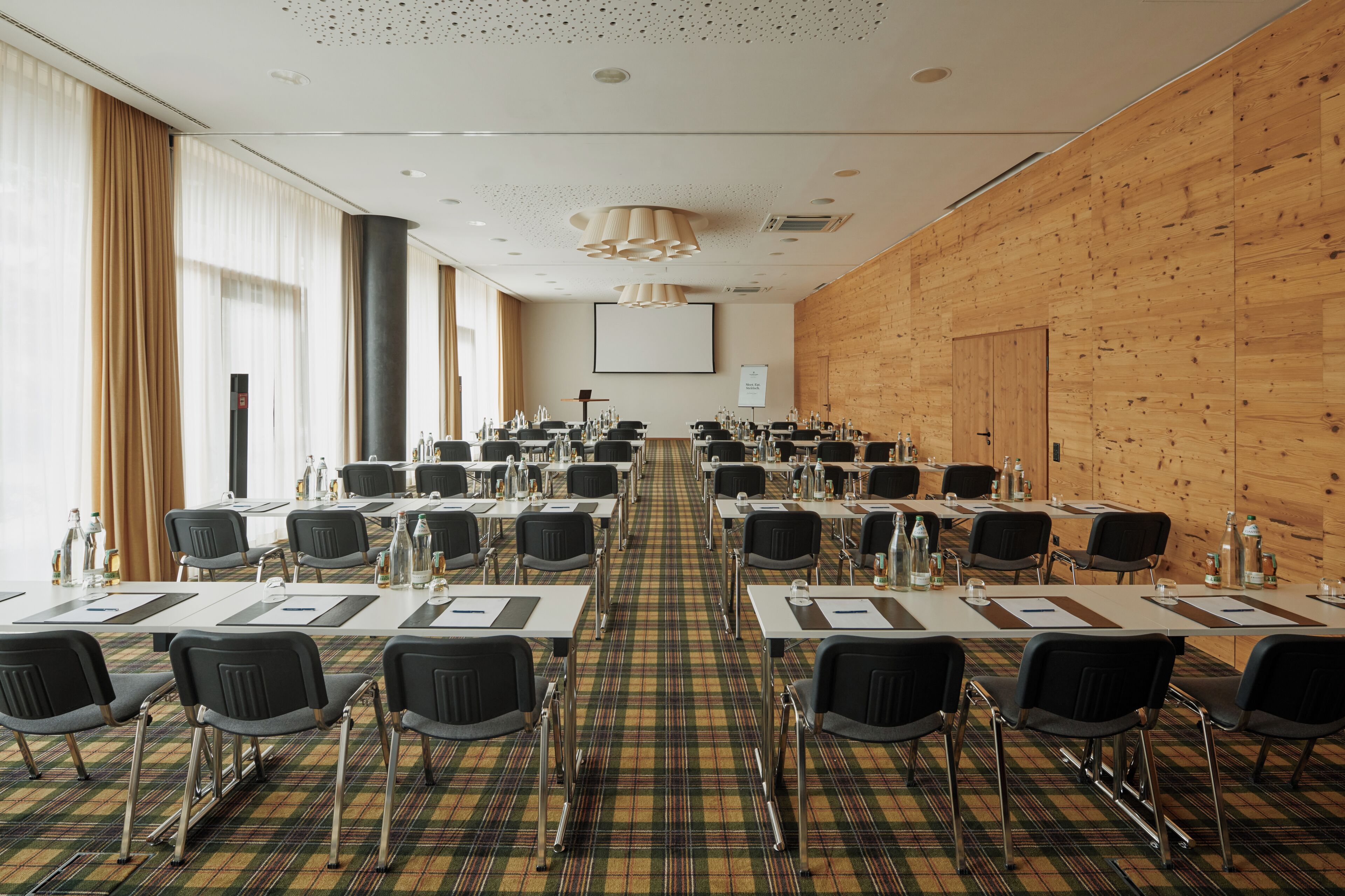 A conference room with a long table, chairs, a projector screen, and a wooden wall.