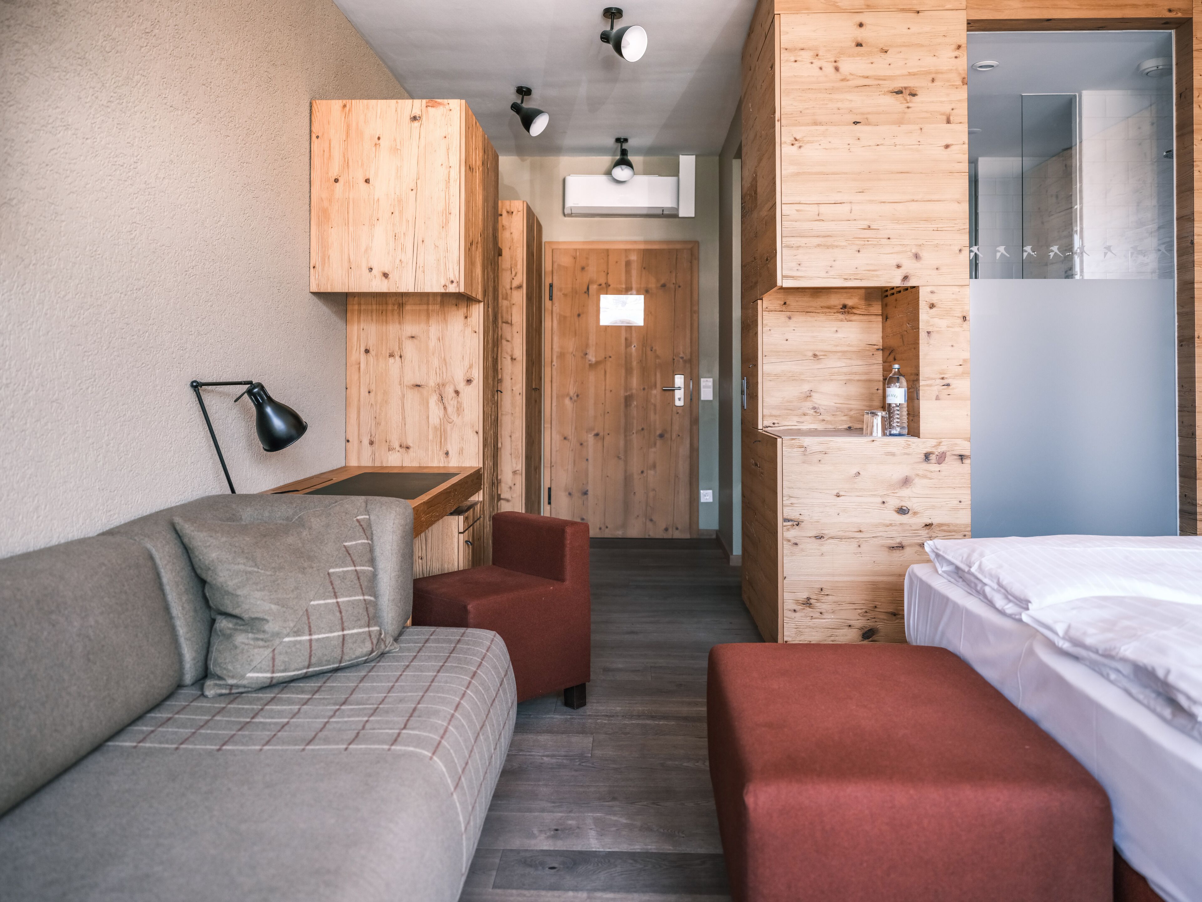 A modern, compact room with a wooden accent wall, a sofa, a red chair, a small table, and a bed.