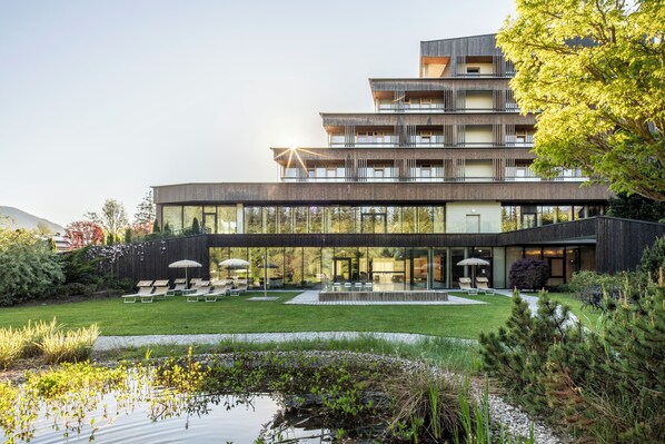 A modern multi-story building with a glass facade, a rooftop terrace with lounge chairs, and a landscaped garden with a pond. - Falkensteiner Hotel Schladming l 4 Star Superior (Schladming)