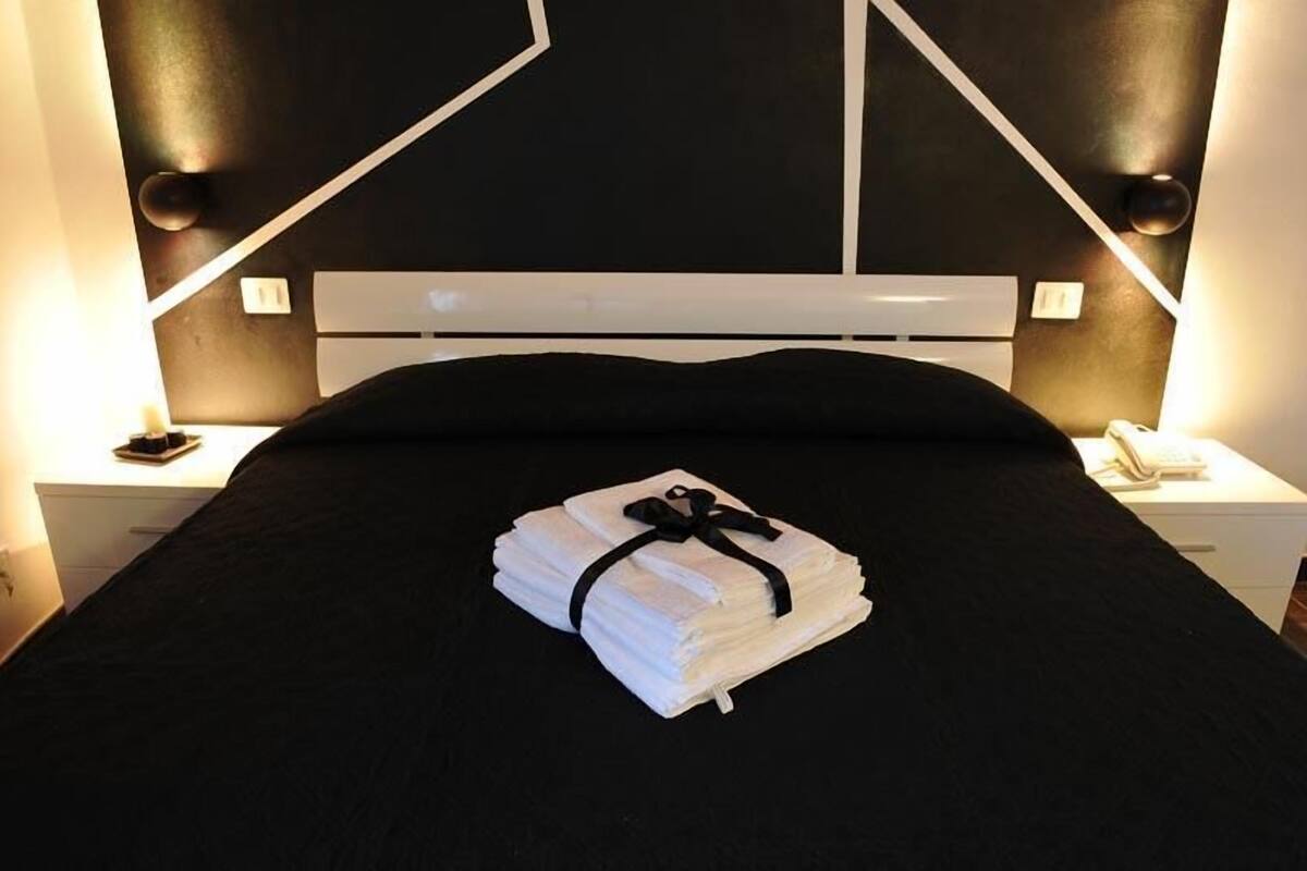 Un lit bien fait, avec une serviette pliée et un ruban noir, une table de chevet blanche avec un téléphone, et un mur orné d’un motif géométrique.