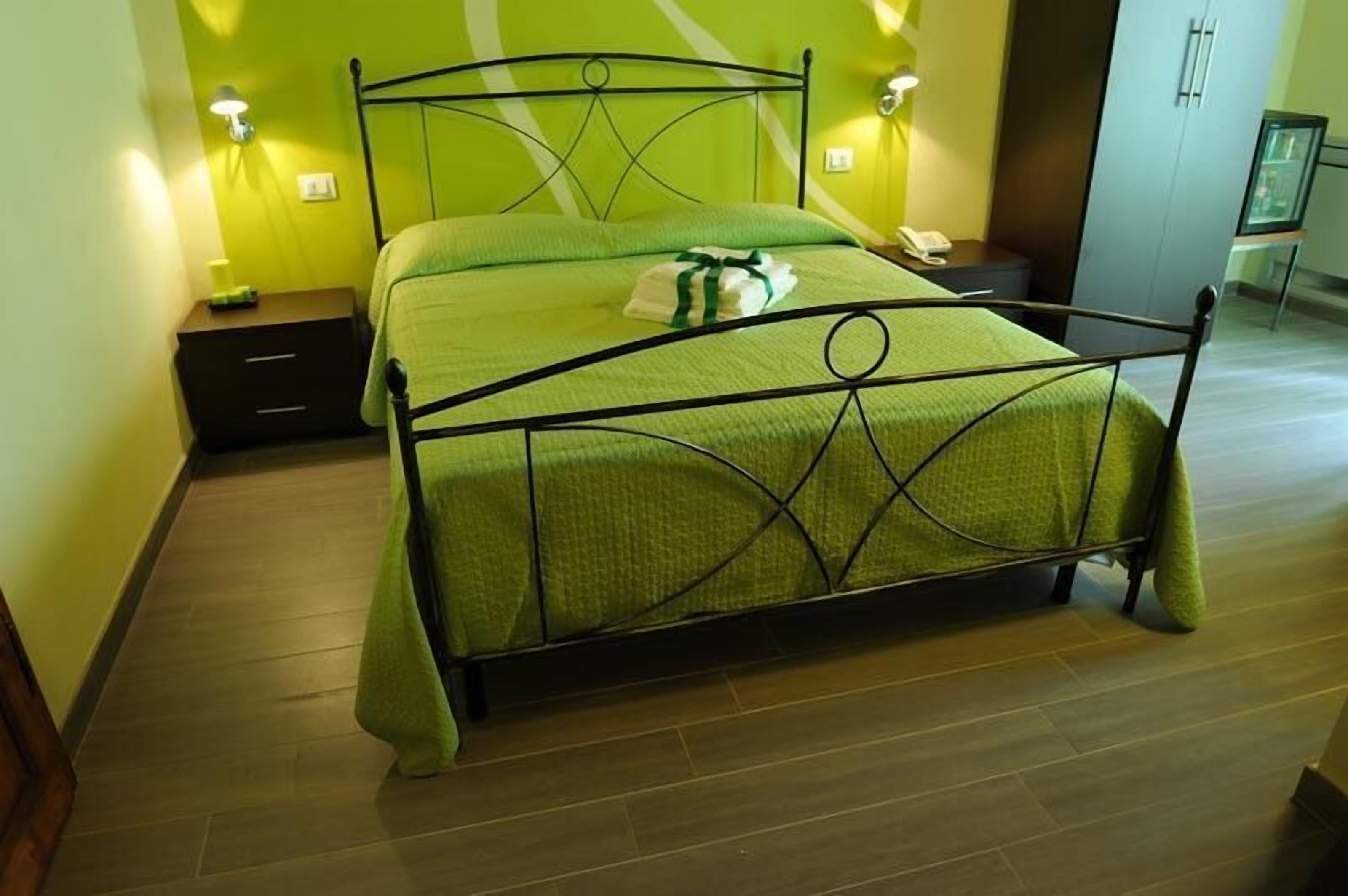 A neatly made bed with a green bedspread and a white towel, a bedside table with a lamp, and a nightstand with a phone.