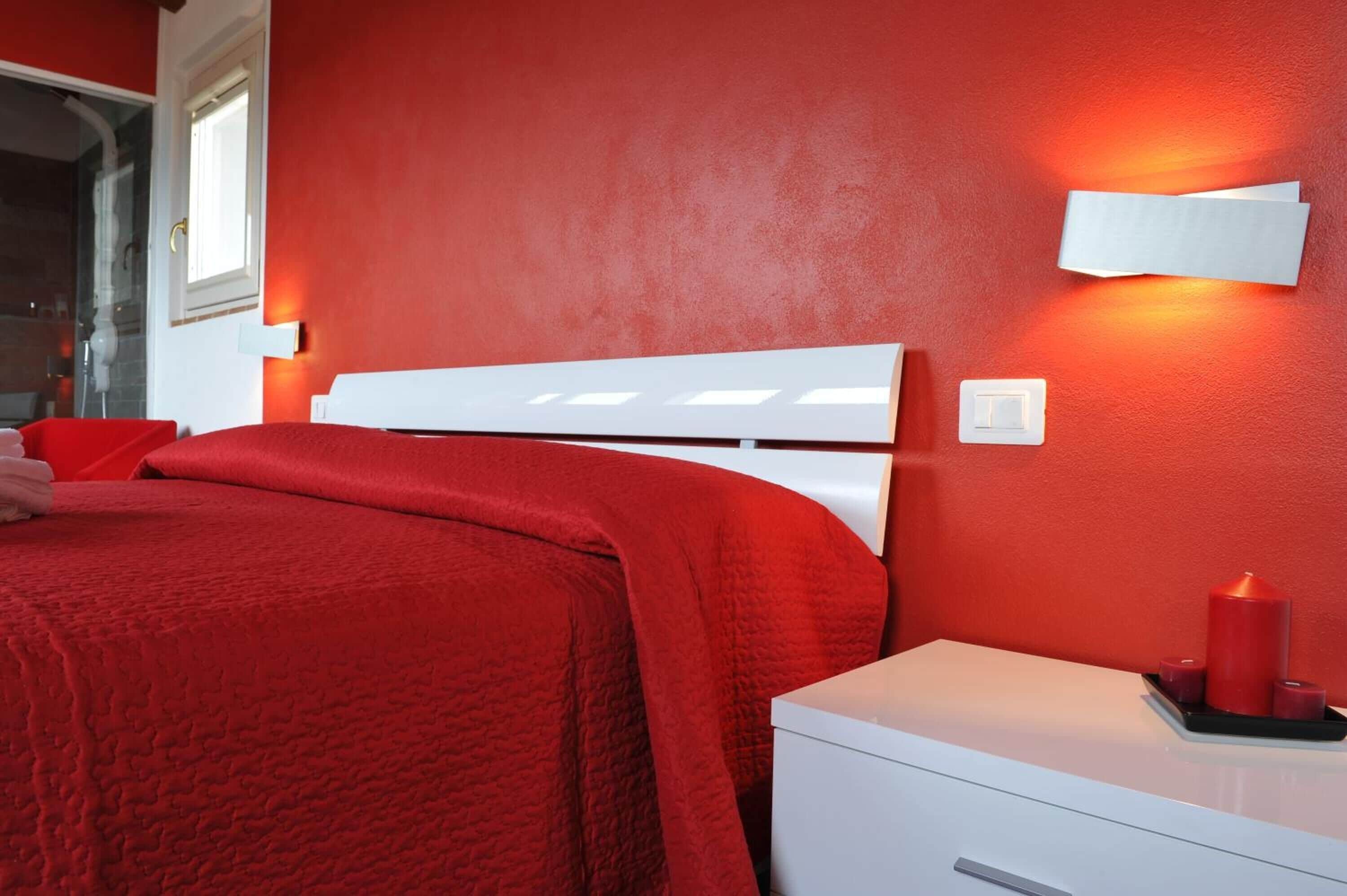 Una habitación con una pared roja, una cama blanca con cabecera, una mesita de noche con una vela roja y un tocador blanco.