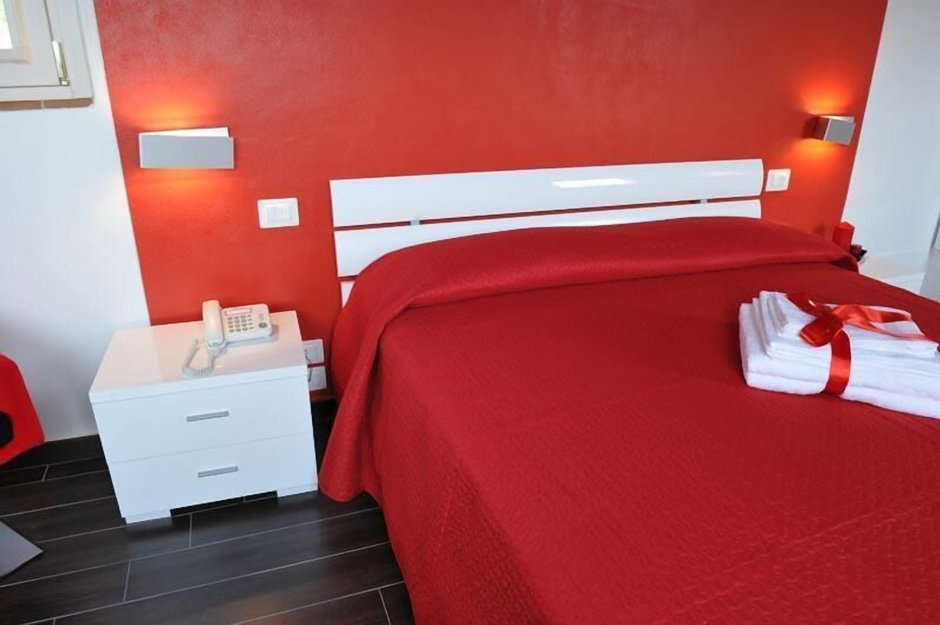 Een hotelkamer met een rood sprei, een witte nachtkastje met telefoon, en een rode stoel.
