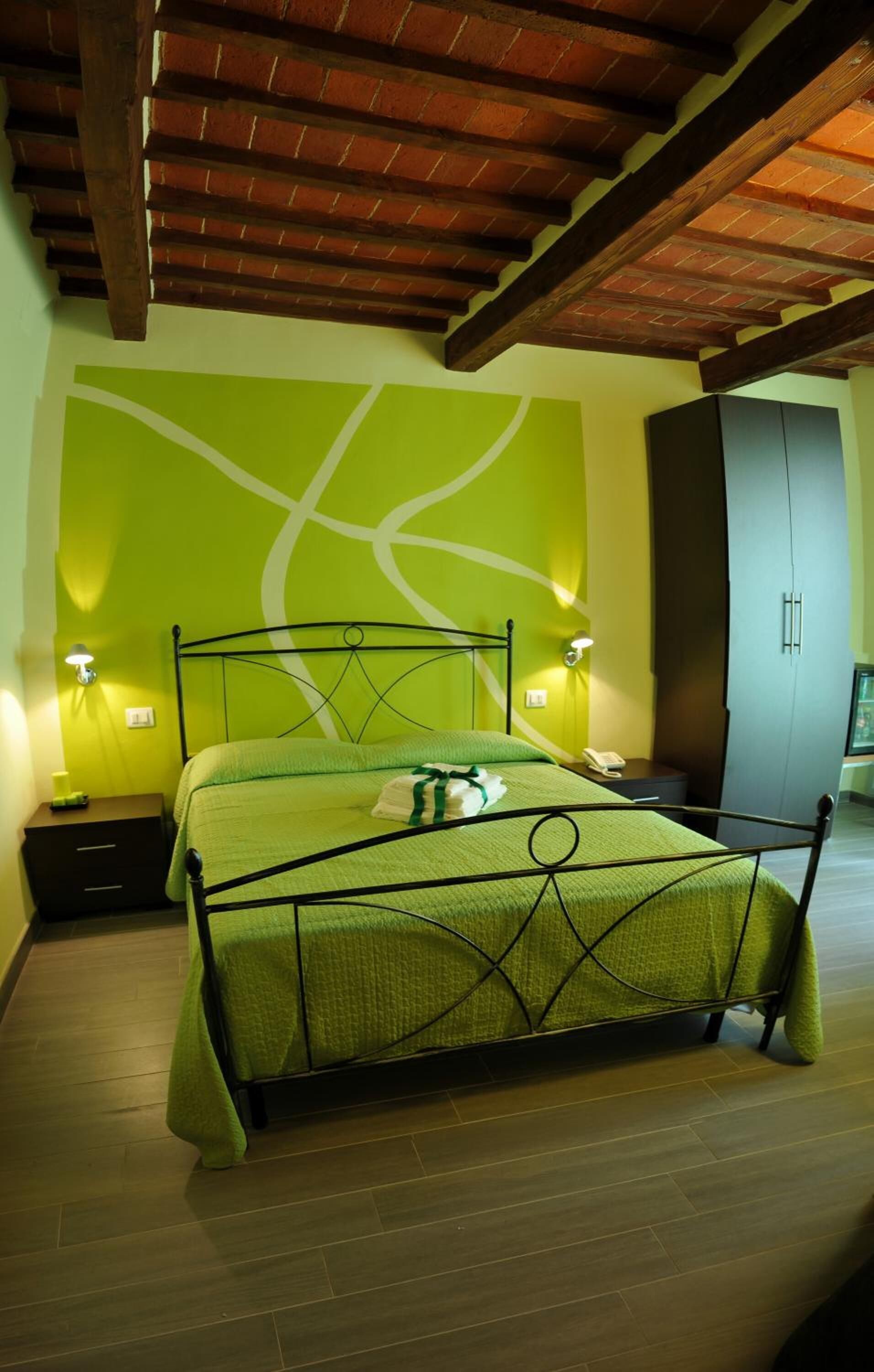 A neatly made bed with a green bedspread, two bedside tables with lamps, a wardrobe, and a wooden ceiling.