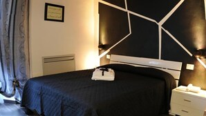 Quarto com cama de cor escura, vigas de madeira no teto, mesa de cabeceira branca e luminária fixada na parede.