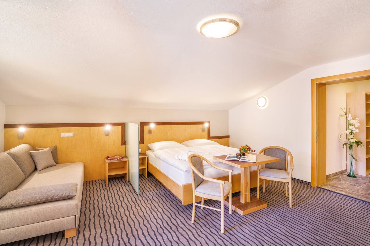 Une chambre d’hôtel comprenant un lit, un canapé, un bureau avec une chaise et une petite table sur laquelle est posé un vase de fleurs.