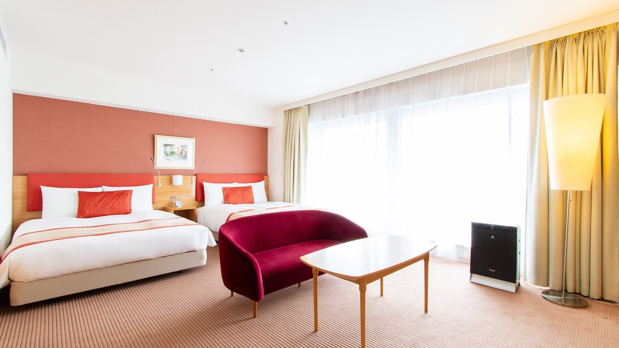 A hotel room with two beds, a red armchair, a small table, a lamp, and a large window with curtains.