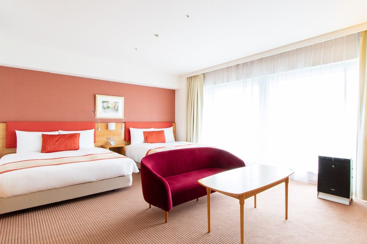A hotel room with two beds, a red armchair, a small table, a lamp, and a large window with curtains.