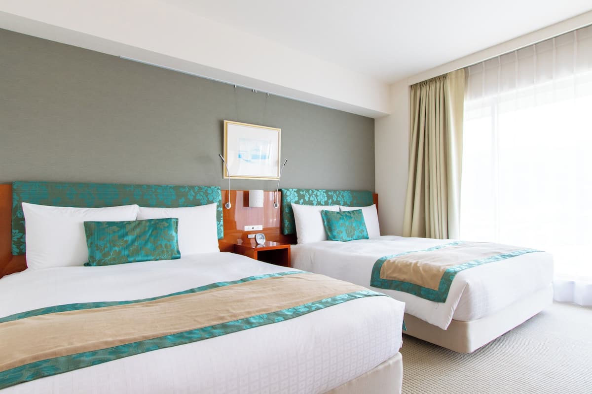 Habitación de hotel con dos camas, un ventanal con cortinas, un cuadro enmarcado en la pared y una mesita de noche de madera.