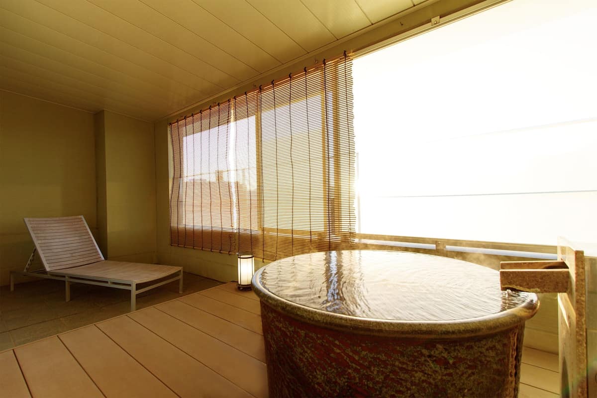 A room with a tatami mat floor, a wooden bed, a round hot tub, and a window with bamboo blinds.