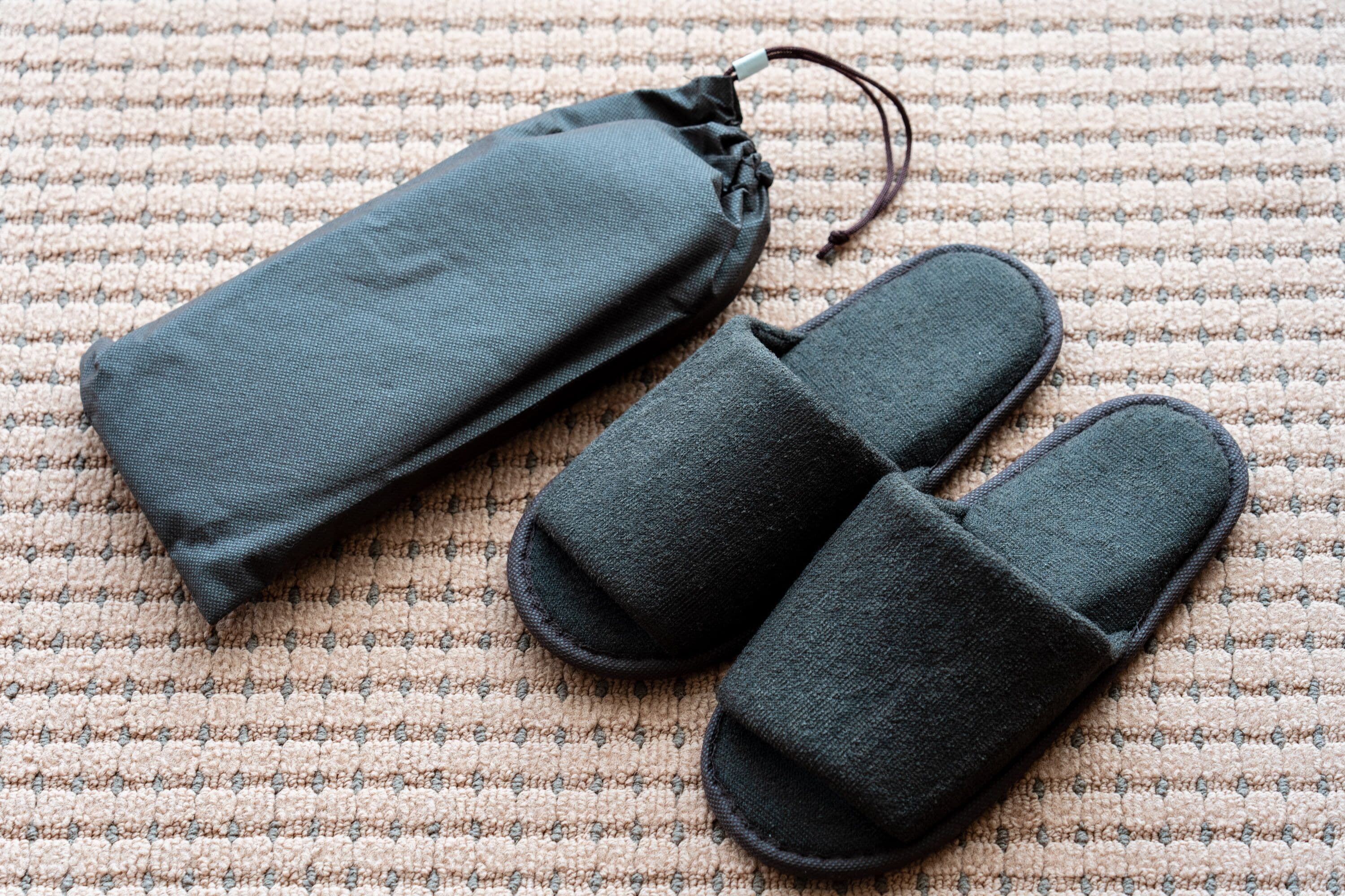 Graue Slipper und eine passende Stofftasche auf einem strukturierten beigen Teppich.