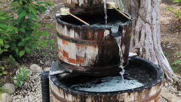 A multi-tiered water fountain made of weathered barrels, set in a garden with trees and pebbles.