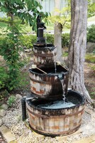 A multi-tiered water fountain made of weathered barrels, set in a garden with trees and pebbles.