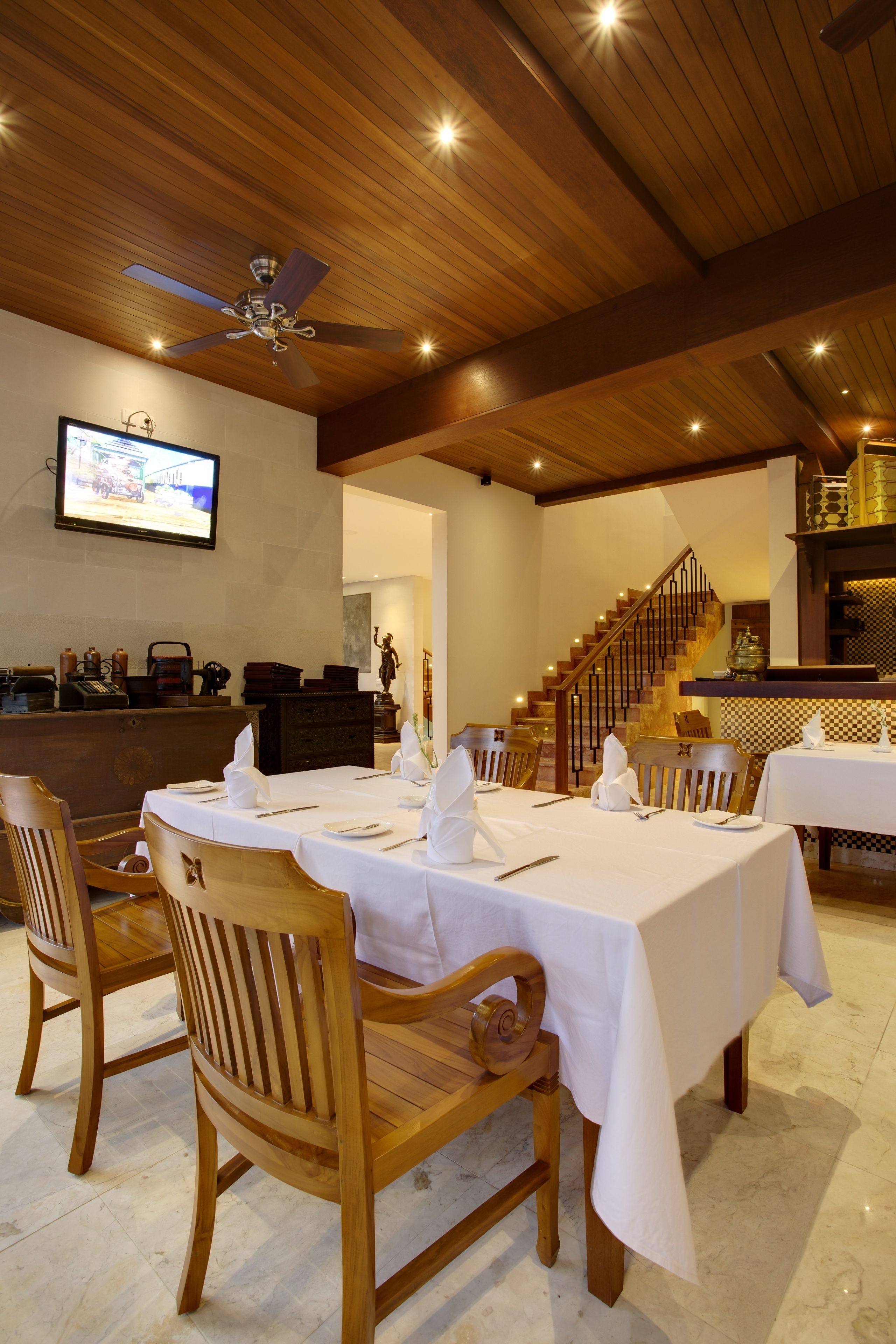 A dining area with wooden tables and chairs, a ceiling fan, a staircase, and a television mounted on the wall.