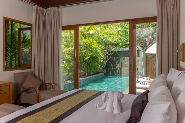 Habitación de hotel con cama, una silla y vista a una piscina exterior rodeada de exuberante vegetación.