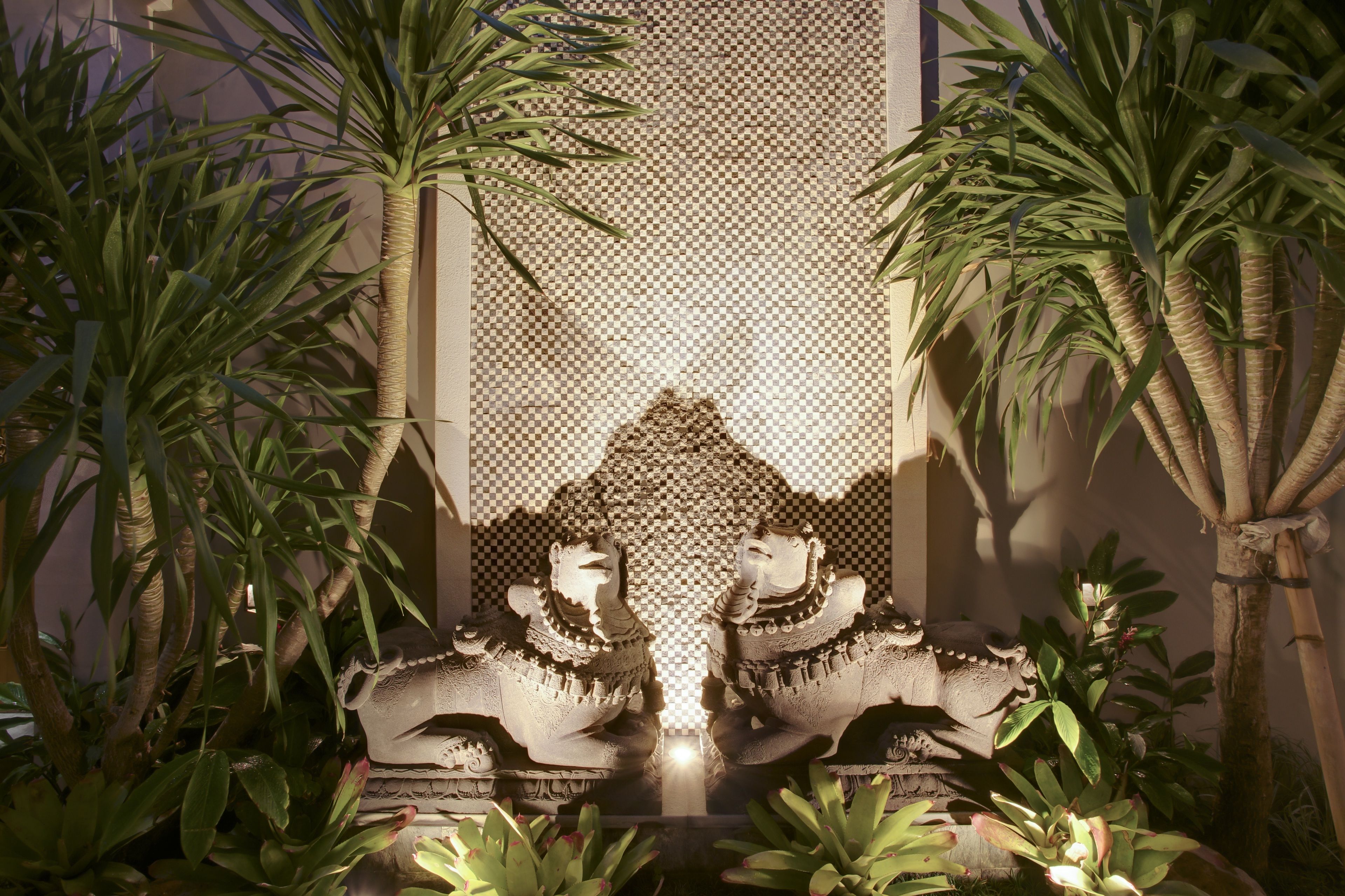 Eine dekorative Steinskulptur mit zwei Figuren in einer Gartenanlage, umgeben von Palmen und anderer Bepflanzung.