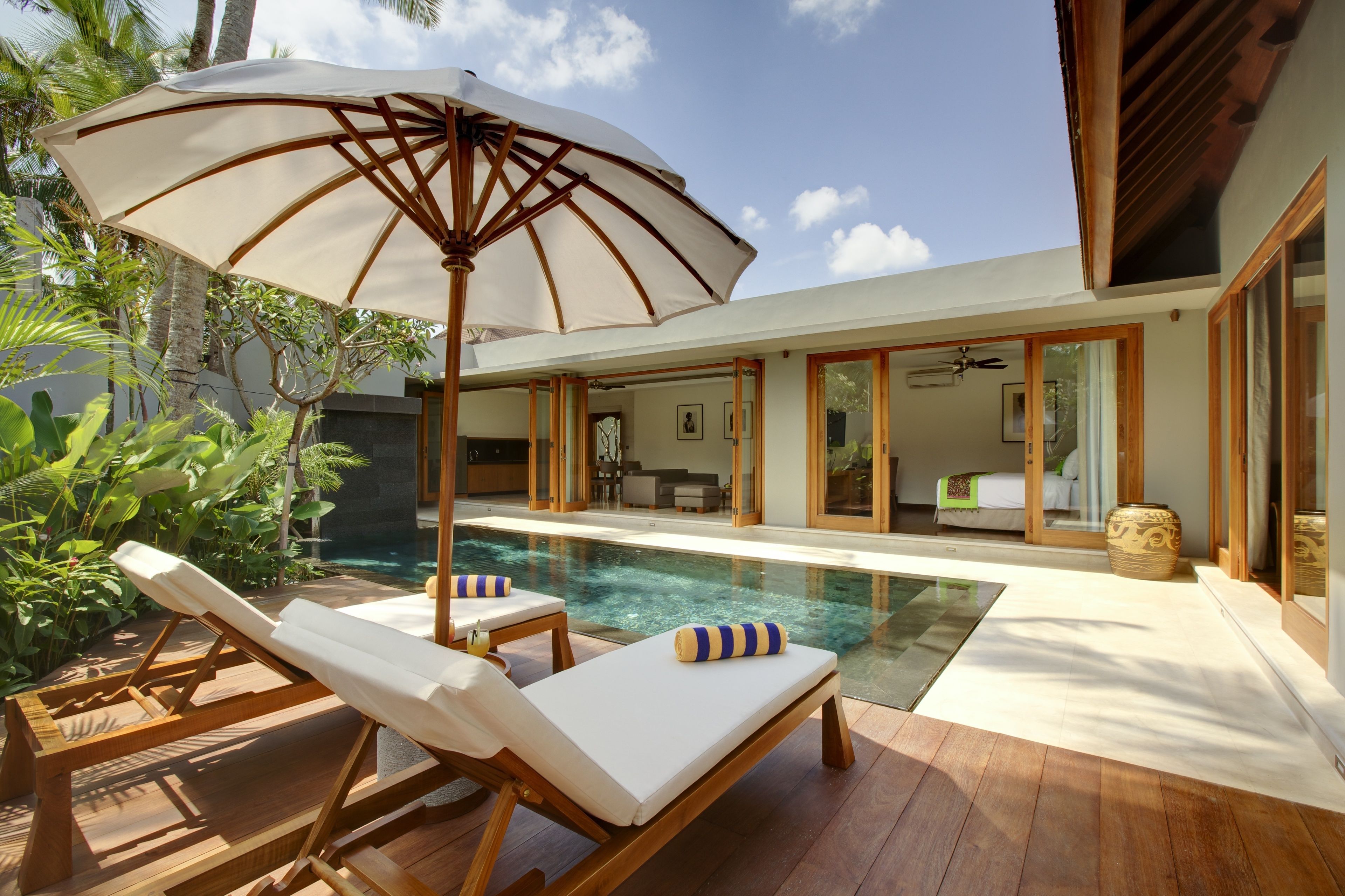 Een modern zwembad met houten ligbedden en een grote parasol, omgeven door tropische planten en een glazen ruimte.