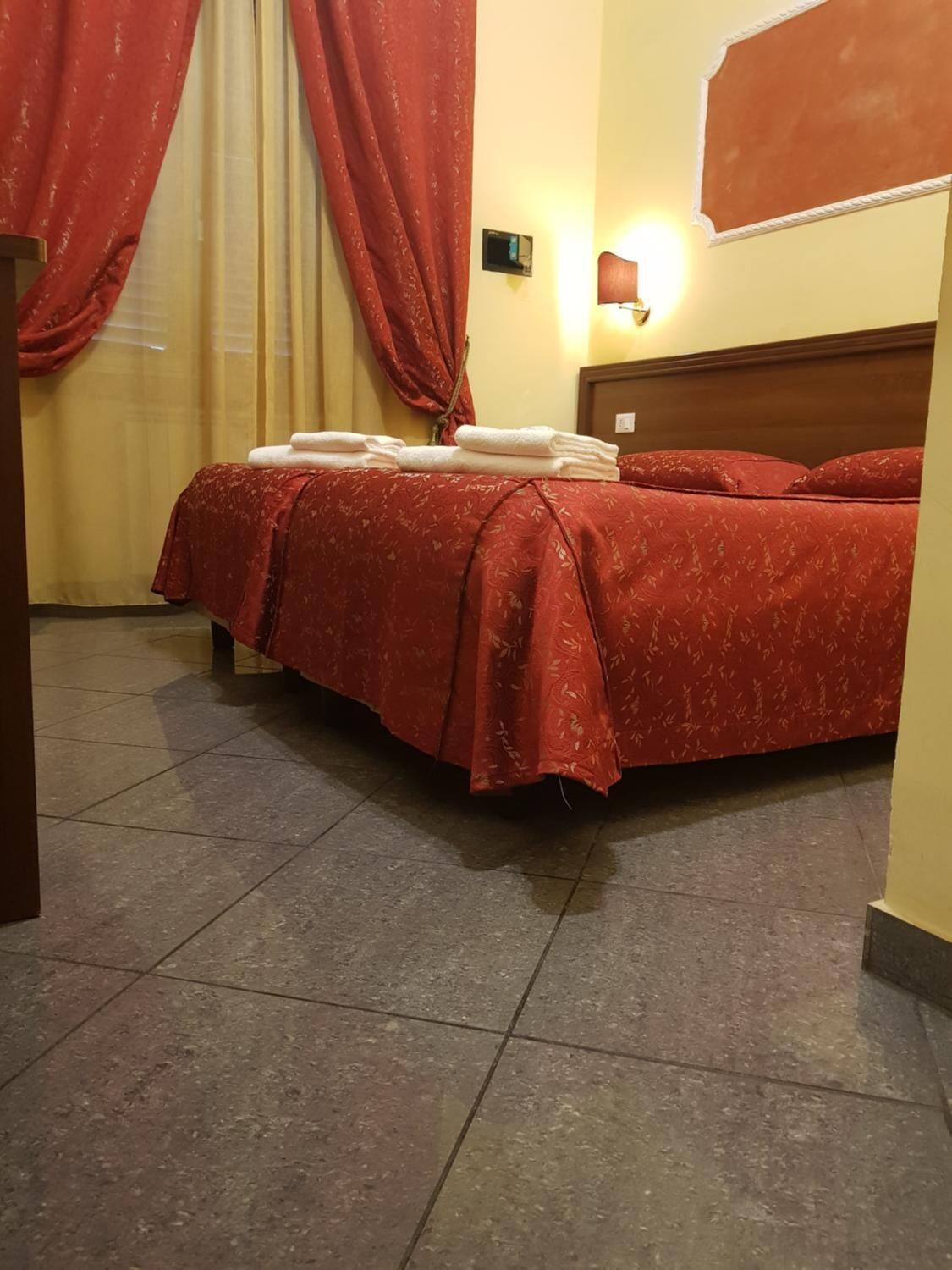 A hotel room with a bed, red curtains, a wall-mounted light, and a tiled floor.