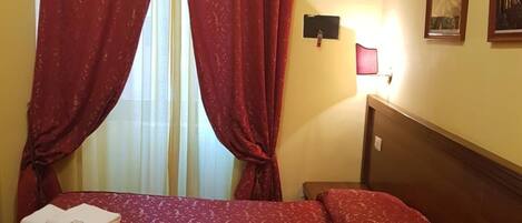 Habitación de hotel con cama, dos toallas, cortinas rojas, mesita de noche, lámpara y dos cuadros en la pared.