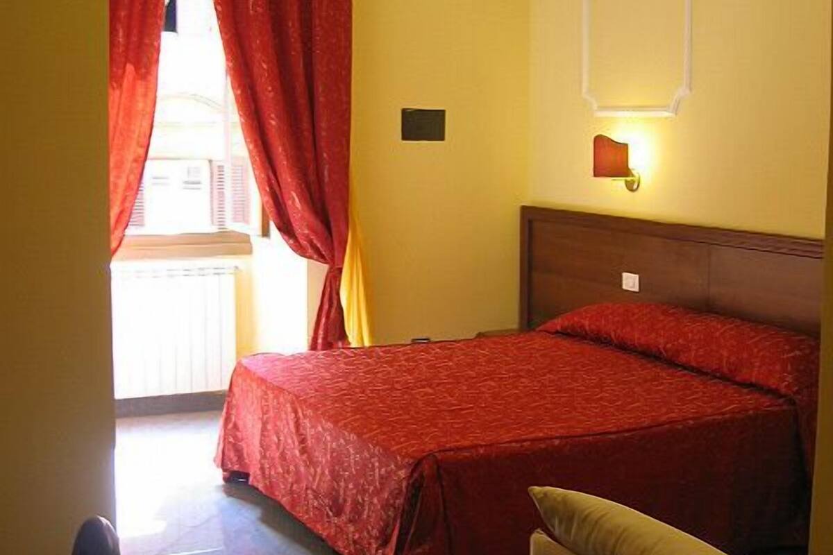 Una habitación de hotel con cama, mesita de noche, lámpara de pared y ventana con cortinas rojas.