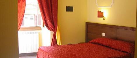 Una habitación de hotel con cama, mesita de noche, lámpara de pared y ventana con cortinas rojas.