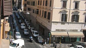 Une vue de rue avec une rangée de voitures garées, un immeuble sur lequel on peut voir une enseigne de restaurant, et des piétons.
