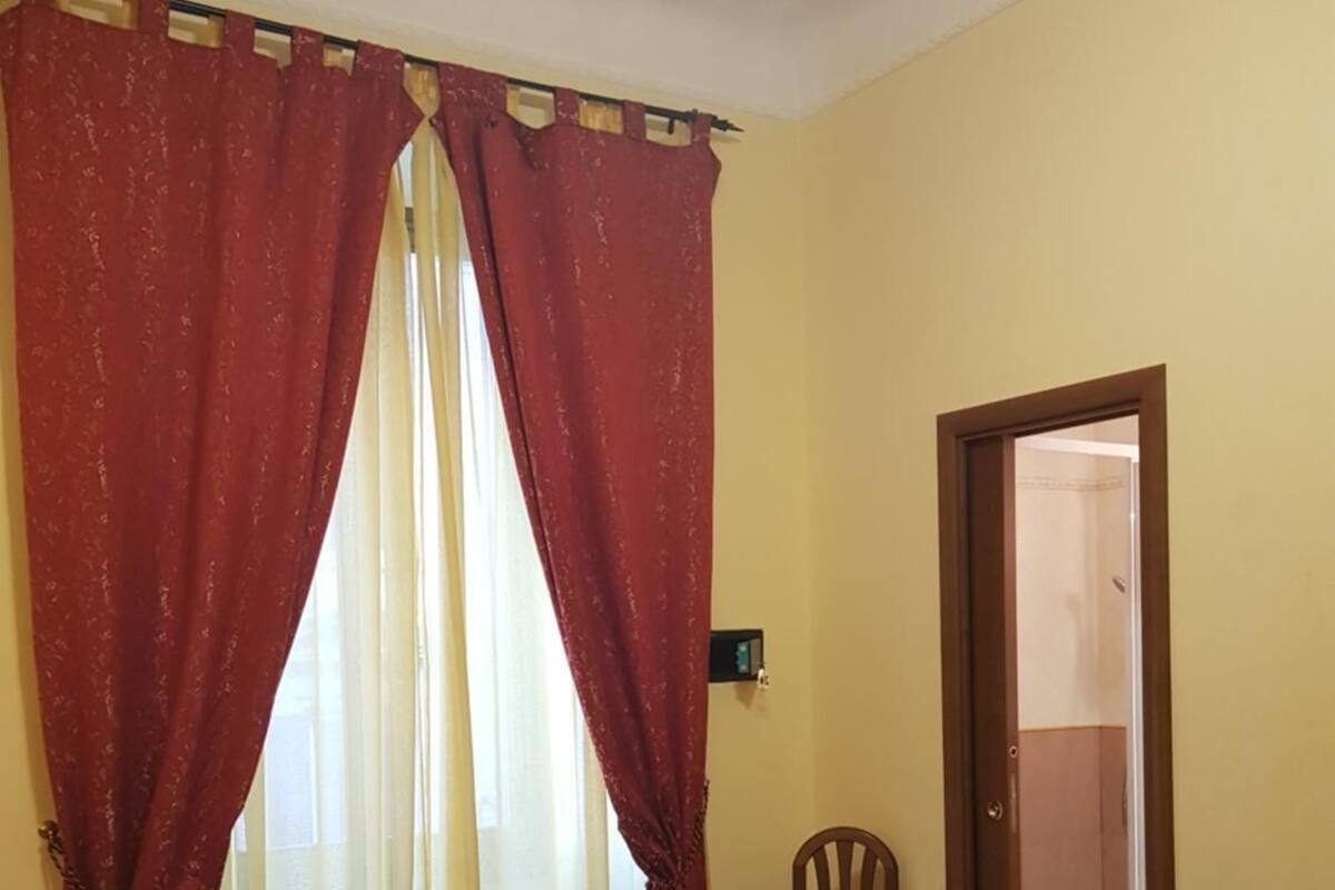 Habitación de hotel con cama, cortinas rojas, una silla y una puerta que da a un baño.