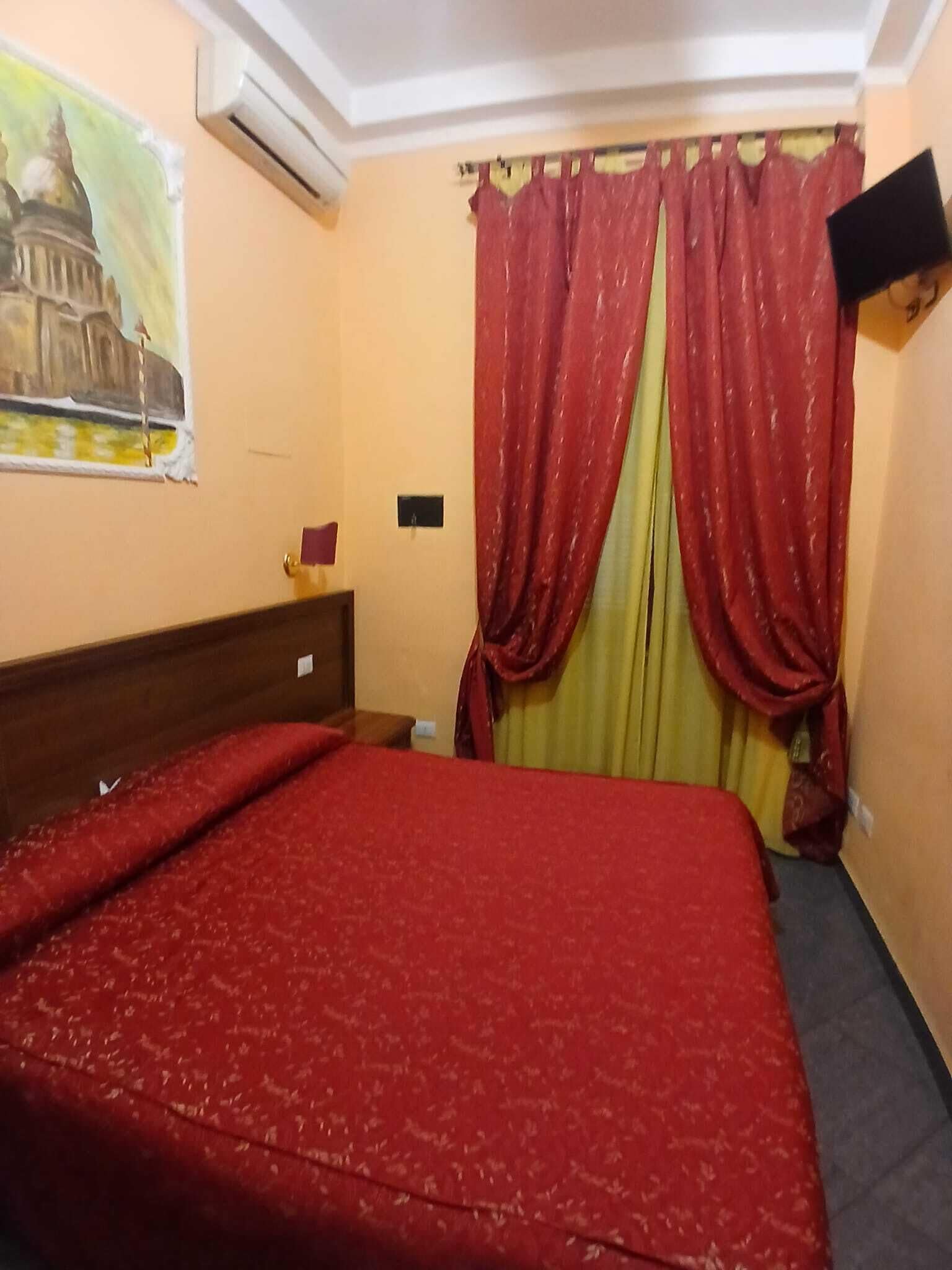 Una habitación de hotel con una cama, un cuadro de un edificio, un televisor en la pared y cortinas rojas.
