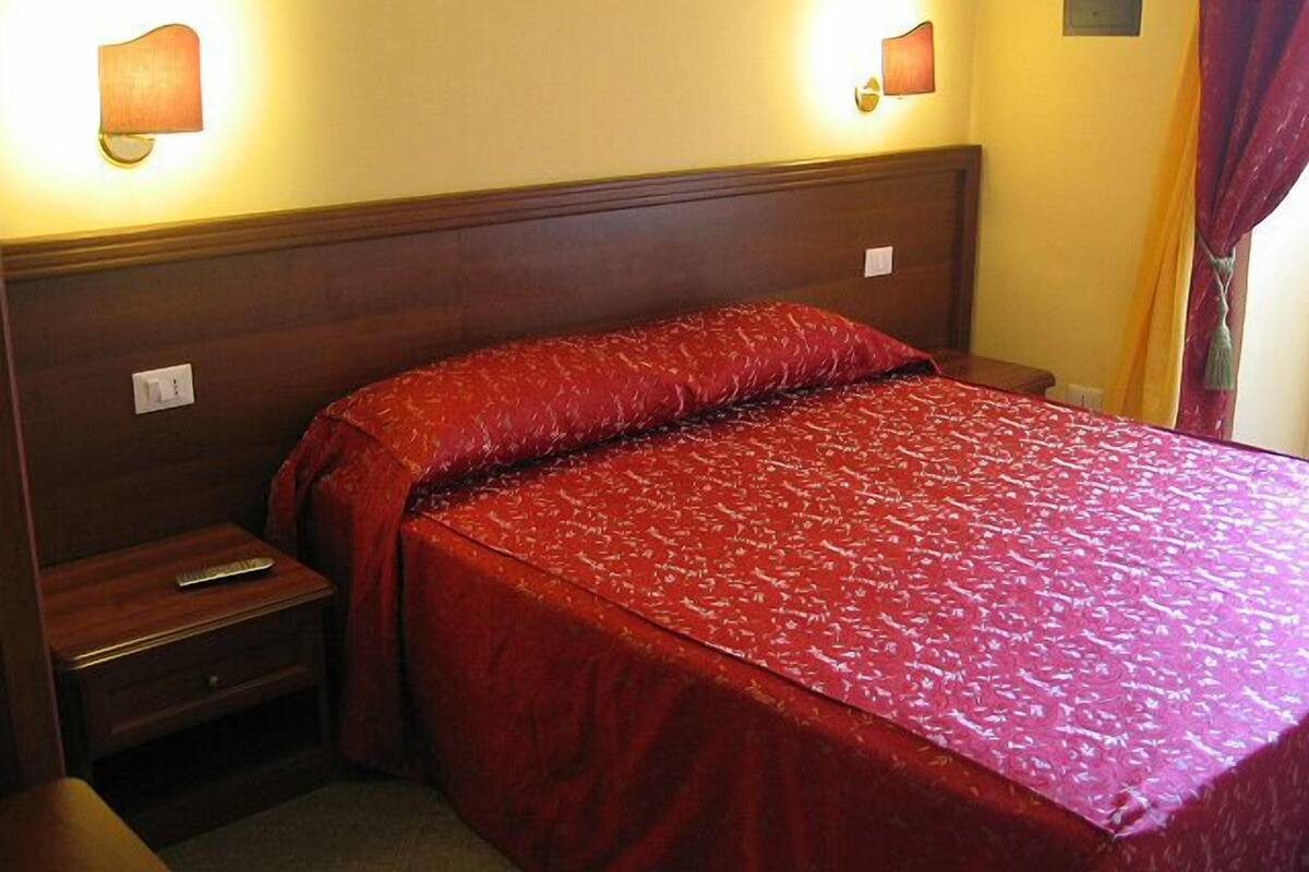 Una habitación de hotel con cabecera de madera, colcha roja y dos lámparas fijadas a la pared.