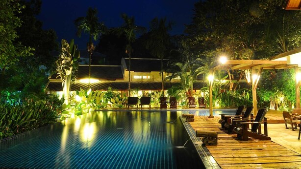 A nighttime pool area with a wooden deck, lounge chairs, and a building in the background. - Lima Bella Resort (Rayong)
